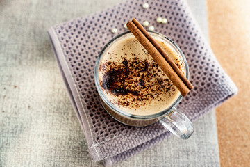 Hot cappuccino with chocolate chips on top of the foam, a cinnamon stick, Golden sugar beads and a sweet waffle next to it. Drink in a transparent Cup on a soft cloth