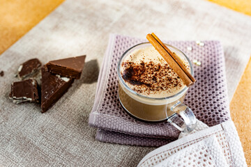 Hot cappuccino with chocolate chips on top of the foam, a cinnamon stick, Golden sugar beads and a sweet waffle next to it. Drink in a transparent Cup on a soft cloth