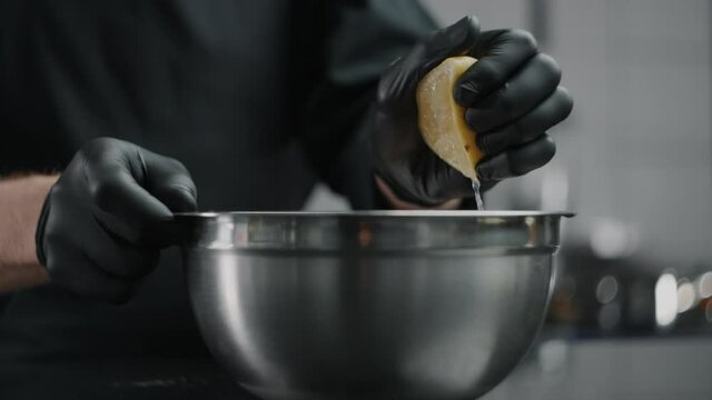 Chef in black gloves squeezes lemon to the metal bowl in slow motion, lemon sprinkles, raw natural ingredients, confectioner chocolatier at work, tabletop video, sour taste, 1080p 240fps Prores HQ