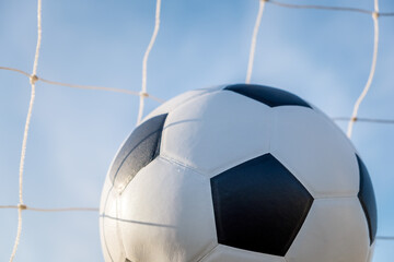 Naklejka premium View of classic leather soccer ball in goal net on blue sky nature background. Traditional black and white football equipment to play competitive game sport championship tournament concept. Copy space