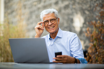 smiling businessman sitting outside with laptop and mobile phone