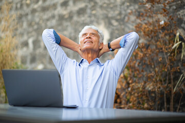 older businessman sitting outside with laptop relaxing with hands behind head