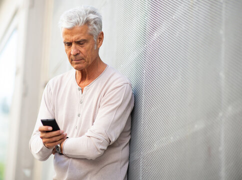 Older Man Leaning Against Wall Looking At Mobile Phone