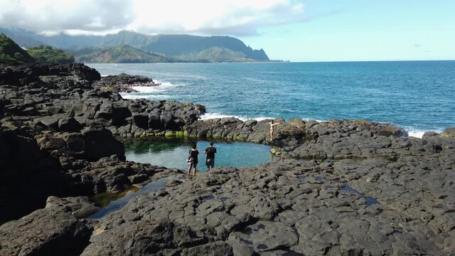 4K Hawaii Kauai static of two family members or friends taking a picture of a woman by Queen's Bath pool with the ocean and mountainous shoreline in the distance with mostly sunny sky