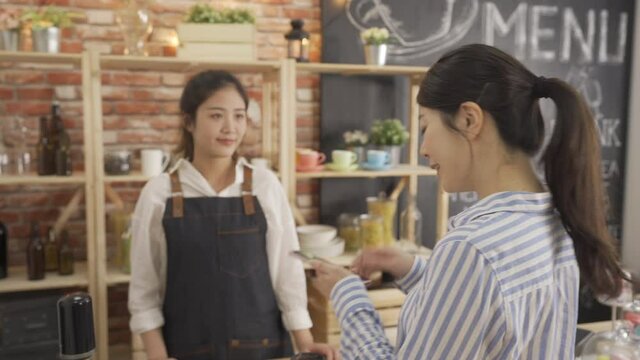 asian female bartender in apron standing in counter give regular customer cup of hot tea every morning daily routine. office lady worker doing smart phone payment in coffee store. waitress waiting.