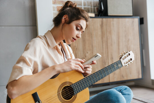 Image of young woman holding cellphone while playing guitar at home