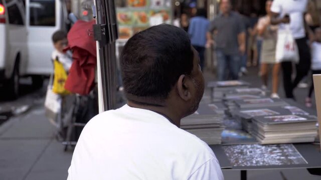 Slow motion of afroamerican black man with white T-shirt sitting on the chair looking around and selling art paintings in New York City. Random people passing by on the busy streets of Manhattan.