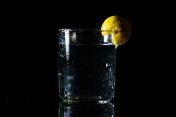 A glass of water placed on a reflective surface in a dark background and a cut lemon on the edge of the glass