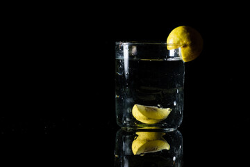 A glass of water placed on a reflective surface in a dark background and a cut lemon inside and on the edge of the glass