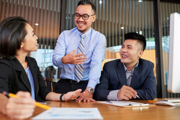 Young adult Asian man standing at desk in board room sharing his ideas to colleagues during business meeting