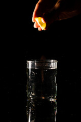 A glass of water placed on a reflective surface in a dark background and a piece of cut lemon is being squeezed from above