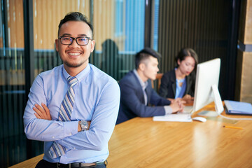 Horizontal medium portrait of confident Asian businessman wearing eyeglasses standing with arms crossed smiling at camera, his colleagues working behind him