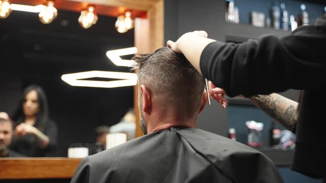 Attractive barber girl cutting man's hair with scissors and comb. Female hair cutter working serving client. Stylish man sitting in vintage barber shop chair. Hairstylist woman doing trendy haircut.