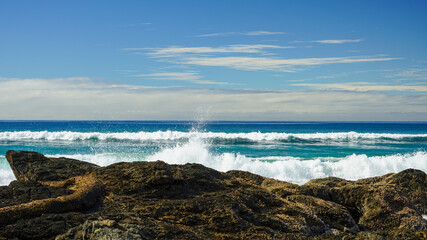 Fototapeta na wymiar Looking out to sea with blue-green waves breaking on rocks in the foreground, and clouds on the horizon, beneath a blue sky. Currumbin, Gold Coast, Queensland, Australia