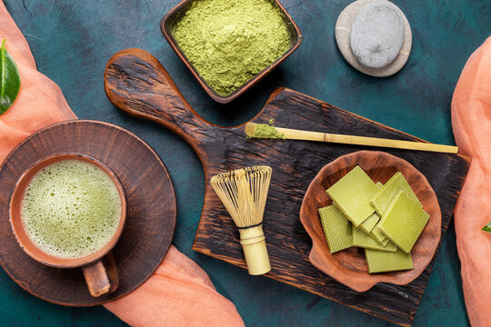 Green matcha tea and powder in brown ceramic cups, matcha chocolate on wooden serving board with coral napkin on emerald backdrop. Matcha tea is healthy detox drink rich of antioxidants. Top view.