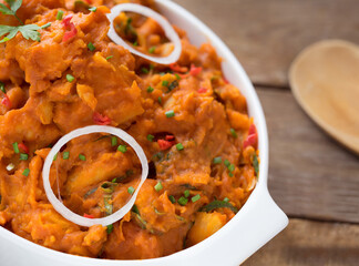 A big white bowl of tasty yam pottage decorated with some green leaf spices, chopped pepper and onion rings placed on a wooden table 