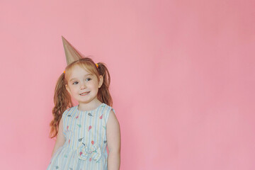 A little cute happy girl in birthday cap smiling with happy face on a pink background. Copy space