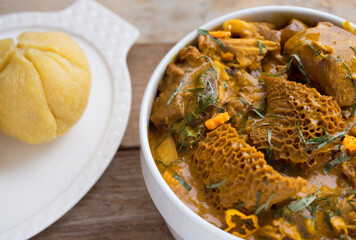 A white bowl of Nigerian dish Garri or eba and Oha soup cooked with assorted meat, beef, dried fish and cow belly meat 'shaki' decorated with green leaf spices and sliced yellow pepper on wood table