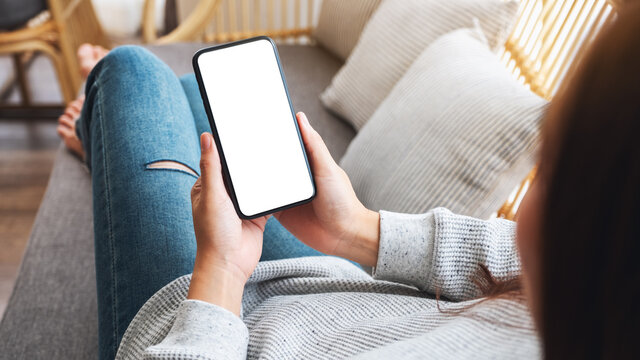 Mockup image of a woman holding mobile phone with blank desktop white screen while lying on a sofa at home
