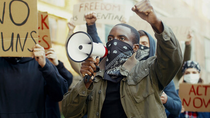 African American young handsome man in scarf on face like mask screaming in megaphone at protest for human rights outdoors. Group of people protesting at street. Strike against violence. - obrazy, fototapety, plakaty