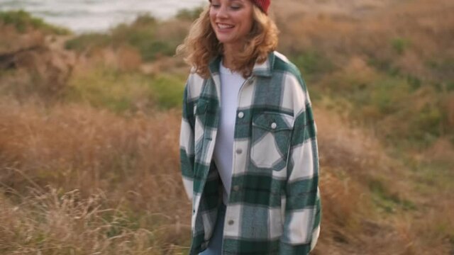 Joyful blonde woman wearing hat and plaid shirt having fun and looking around while walking outdoors