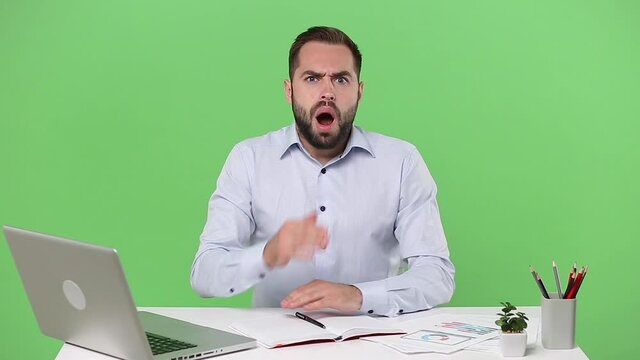 Young shocked surprised bearded business man in light shirt sit work at desk with pc laptop computer spreading hands isolated on green background studio Achievement business career lifestyle concept