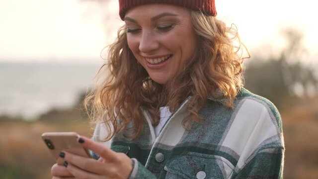Happy blonde woman wearing hat and plaid shirt using smartphone outdoors