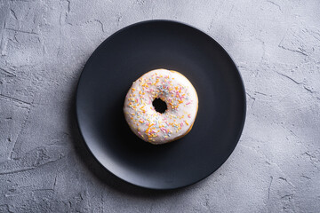 Vanilla donut with sprinkles on black plate, sweet glazed dessert food on concrete textured background, top view