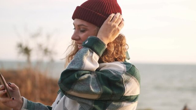 Side view of Happy blonde woman wearing hat and plaid shirt making selfie on smartphone and looking away outdoors
