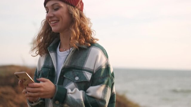 Side view of Joyful blonde woman wearing hat and plaid shirt using smartphone and looking around while walking outdoors