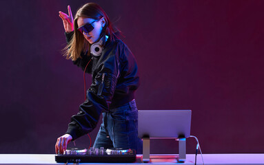 Stylish DJ girl with headphones and glasses on the bomber jacket mixes the music at the party and neon lights.