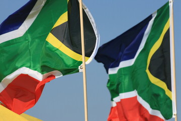 A couple of colourful South African flags blow in the wind whilst mounted onto thin stick poles set against a vivid cloudless blue sky taken during the summer season. London, England