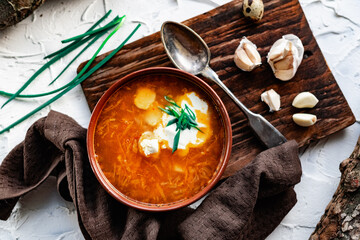 A portion of red borscht in a brown plate, next to an old wooden Board garlic and quail egg, green leeks and driftwood. White sour cream in the soup, next to an old Polish spoon