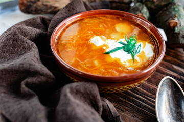 A portion of red borscht in a brown plate, next to an old wooden Board garlic and quail egg, green leeks and driftwood. White sour cream in the soup, next to an old Polish spoon