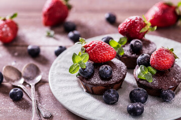 Chocolate cakes (cupcakes) are on the table. Cupcakes decorated  blueberries, strawberries and green leaves mint on top. Beautiful and elegant serving with white dishes