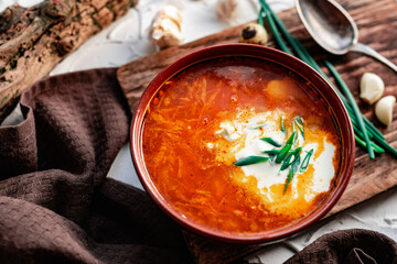 A portion of red borscht in a brown plate, next to an old wooden Board garlic and quail egg, green leeks and driftwood. White sour cream in the soup, next to an old Polish spoon