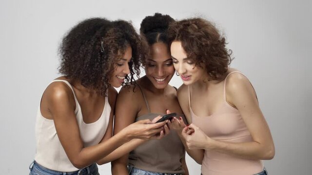 Three happy multi-ethnic women are watching the picture on the smartphone while communicating with each other isolated over white background