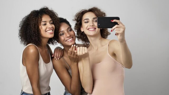 Three beautiful multi-ethnic women are taking selfie-photo on the smartphone isolated over white background