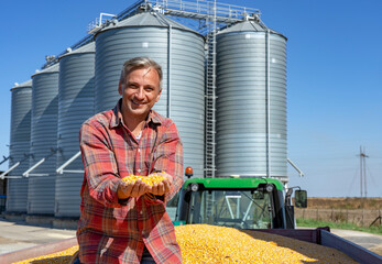 Happy Farmer Showing Freshly Harvested Corn Grains Against Grain Silos © RGtimeline