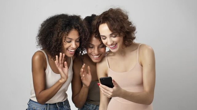 Three smiling multi-ethnic women are talking with video-connection using smartphone isolated over white background