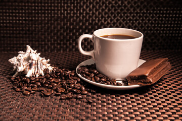 A cup of coffee with a chocolate bar, scattered coffee beans and a shell.
