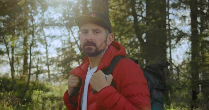 Portrait of serious contemplative young man, travel blogger with adventure backpack and red puffy jacket look into camera. Tourist or hiker walk in sunny summer forest in Norway or Canada
