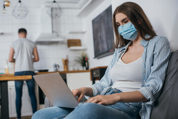 selective focus of young freelancer in medical mask working on laptop in kitchen near boyfriend on background