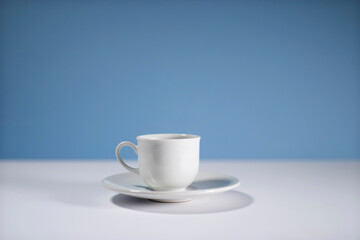 White cup of morning espresso on a stone surface of a table with reflections opposite blue background.