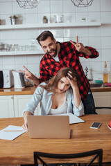 cheerful man fooling around near exhausted girlfriend sitting near laptop and notebook in kitchen