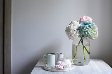 A bouquet of multi-colored hydrangeas in a five-liter jar on a blue wall background. A tray with mugs, milkman and pink marshmallows.