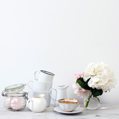 Ball-shaped white hydrangea in a round glass vase, set of light dishes, milk jug, porcelain and enameled cups, a jug for a greeting card on a white background.