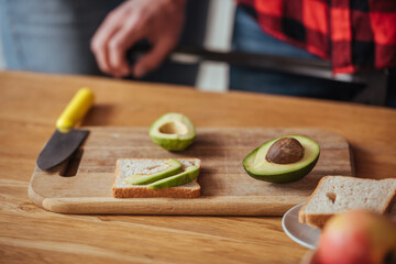 cropped view of man near chopping board with bread, knife and halves of fresh avocado
