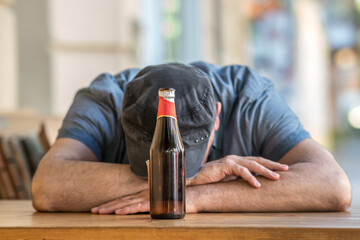 Pensive and sad man with bowed head sitting holding a bottle of beer outside a bar. Selective focus
