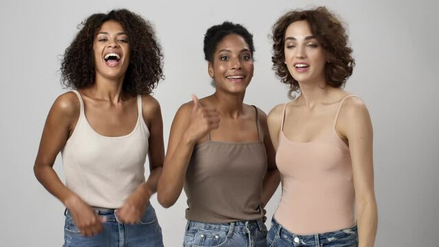 Three happy multi-ethnic women are showing thumb-up gesture standing isolated over white background